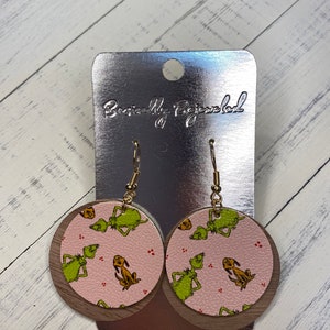 May include: A pair of round, pink and brown earrings with a green and yellow print of the Grinch and Max the dog. The earrings are on a silver card with the text "Beautifully Bejeweled".