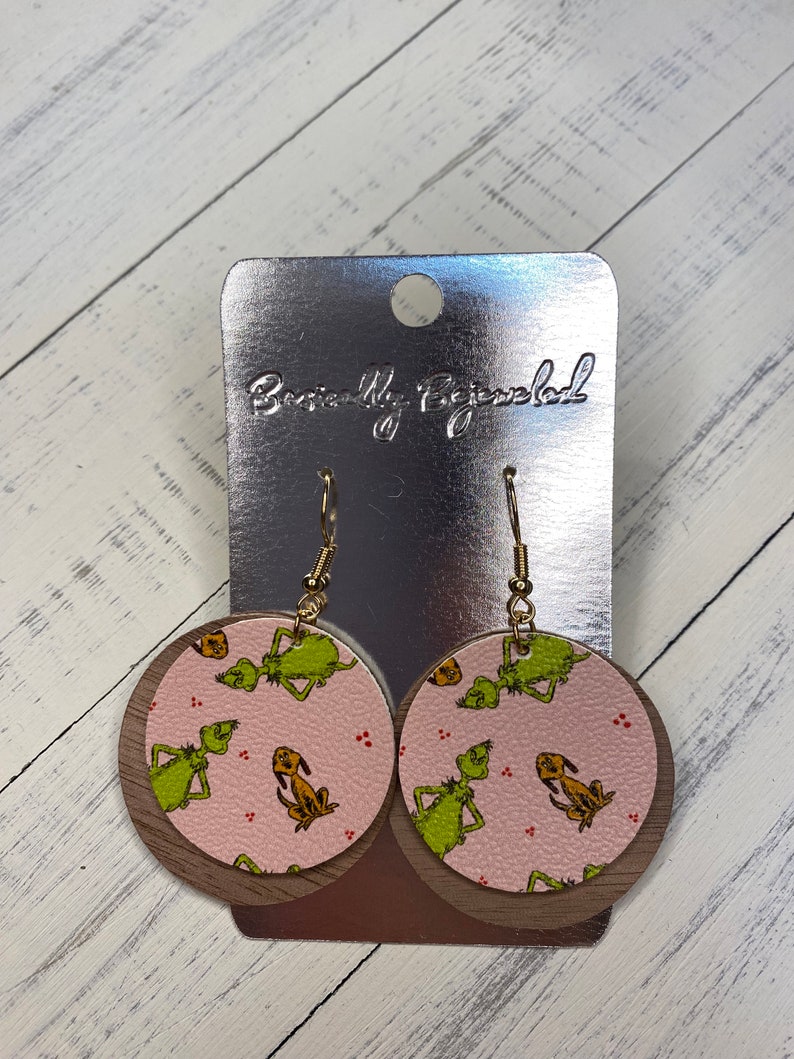 May include: A pair of round, pink and brown earrings with a green and yellow print of the Grinch and Max the dog. The earrings are on a silver card with the text "Beautifully Bejeweled".