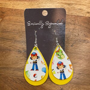 Puede incluir: Un par de pendientes amarillos en forma de l&aacute;grima con un fondo blanco que presenta un personaje de dibujos animados con un sombrero rojo y una camisa azul. Los pendientes est&aacute;n en una tarjeta plateada con el texto "Beautifully Blinged".
