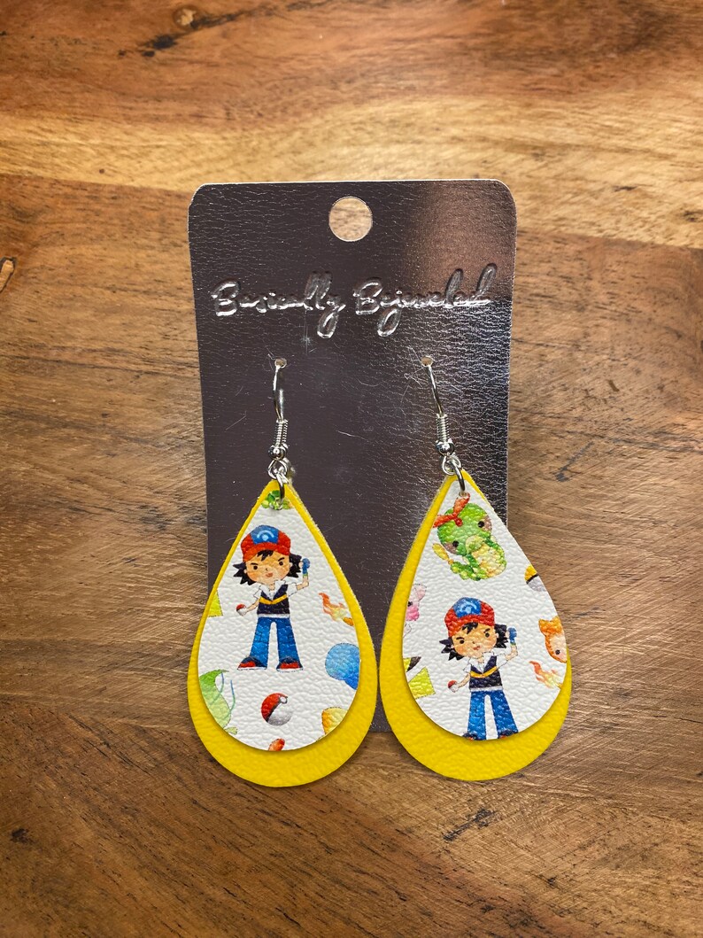 Puede incluir: Un par de pendientes amarillos en forma de l&aacute;grima con un fondo blanco que presenta un personaje de dibujos animados con un sombrero rojo y una camisa azul. Los pendientes est&aacute;n en una tarjeta plateada con el texto "Beautifully Blinged".
