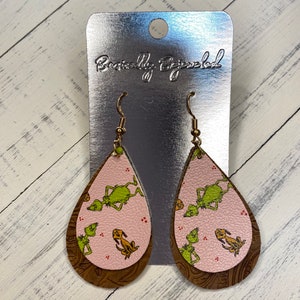 May include: A pair of dangle earrings with a pink and brown leather teardrop design. The teardrop features a green and yellow print of the Grinch and Max from the movie How the Grinch Stole Christmas.