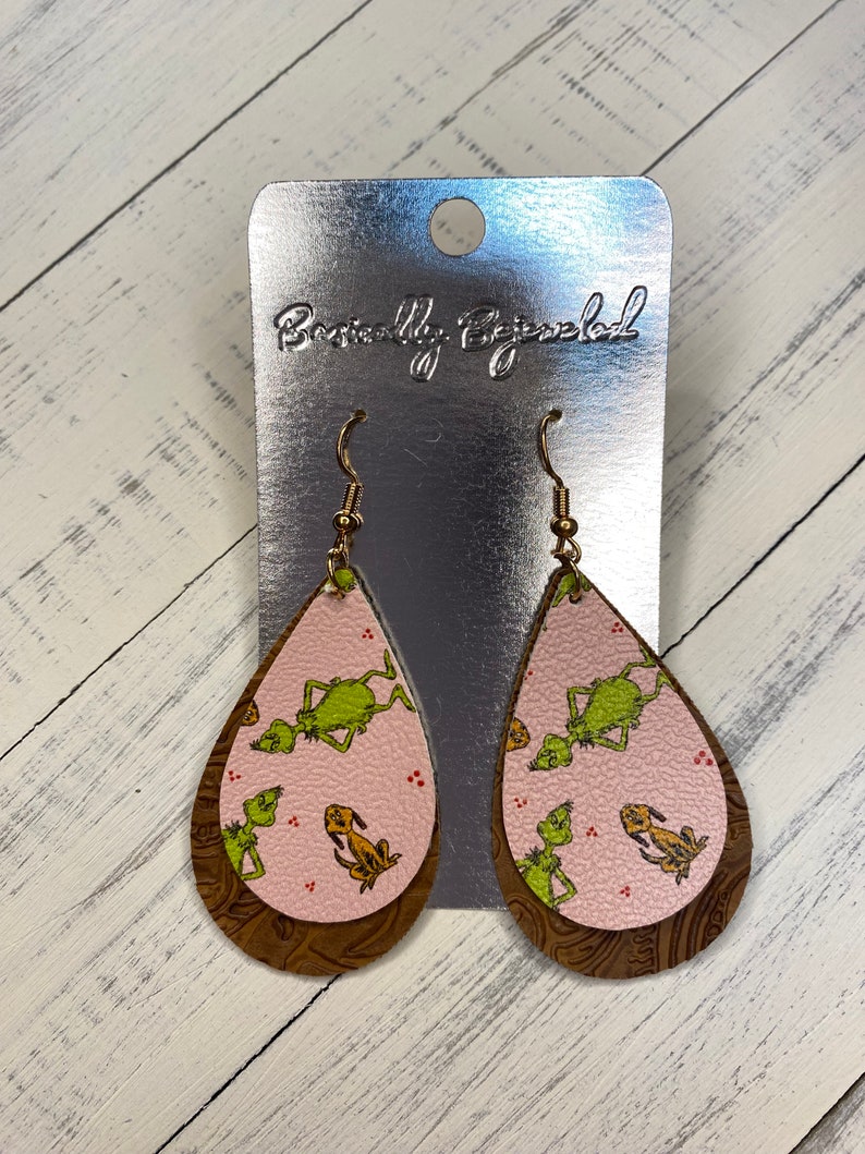 May include: A pair of dangle earrings with a pink and brown leather teardrop design. The teardrop features a green and yellow print of the Grinch and Max from the movie How the Grinch Stole Christmas.