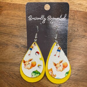 Puede incluir: Un par de pendientes amarillos en forma de l&aacute;grima con un fondo blanco que presenta un personaje de dibujos animados y un dise&ntilde;o de Pok&eacute; Ball. Los pendientes est&aacute;n en una tarjeta plateada con el texto "Beautifully Blessed".