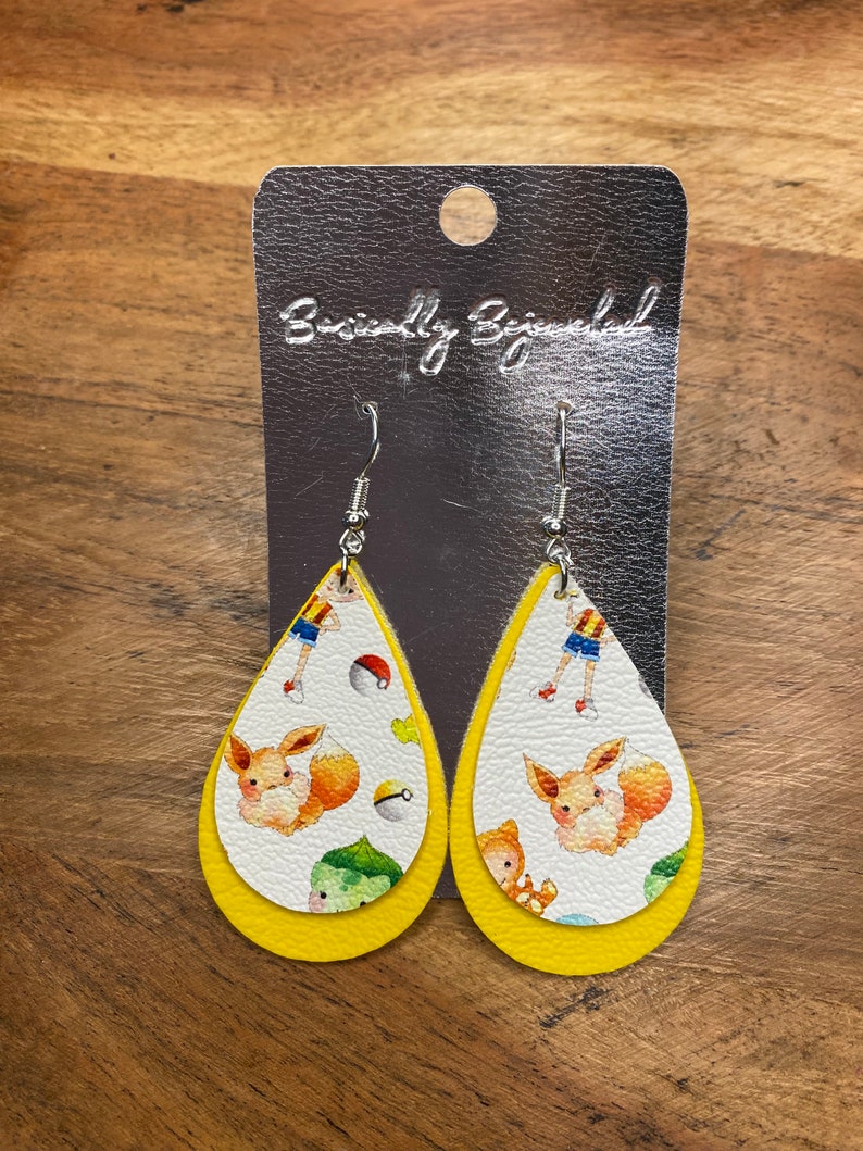 Puede incluir: Un par de pendientes amarillos en forma de l&aacute;grima con un fondo blanco que presenta un personaje de dibujos animados y un dise&ntilde;o de Pok&eacute; Ball. Los pendientes est&aacute;n en una tarjeta plateada con el texto "Beautifully Blessed".