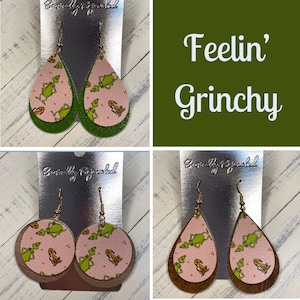 May include: Two pairs of dangle earrings featuring a Grinch pattern on pink and green leather. The earrings are labeled "Beautifully Fringed". The text "Feelin' Grinchy" is displayed on a green background.