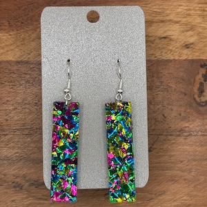 May include: A pair of dangle earrings with a rectangular shape. The earrings are made of a glittery material with a mix of green, blue, pink, and gold colors.