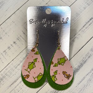 May include: Green and pink dangle earrings with a Grinch pattern. The earrings are on a silver card with the text "Beautifully Bejeweled".