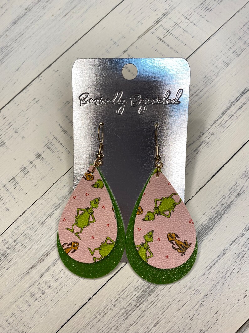 May include: Green and pink dangle earrings with a Grinch pattern. The earrings are on a silver card with the text "Beautifully Bejeweled".