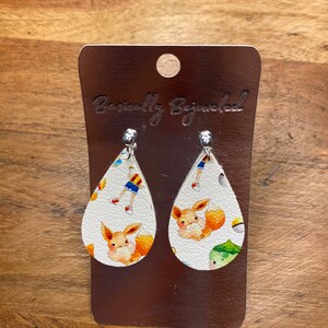 Puede incluir: Un par de pendientes blancos en forma de l&aacute;grima con un estampado de personaje de Pokemon. Los pendientes presentan un peque&ntilde;o Pokemon naranja parecido a un zorro sobre un fondo blanco. Los pendientes est&aacute;n en una tarjeta plateada con el texto "Basically Bejeweled".