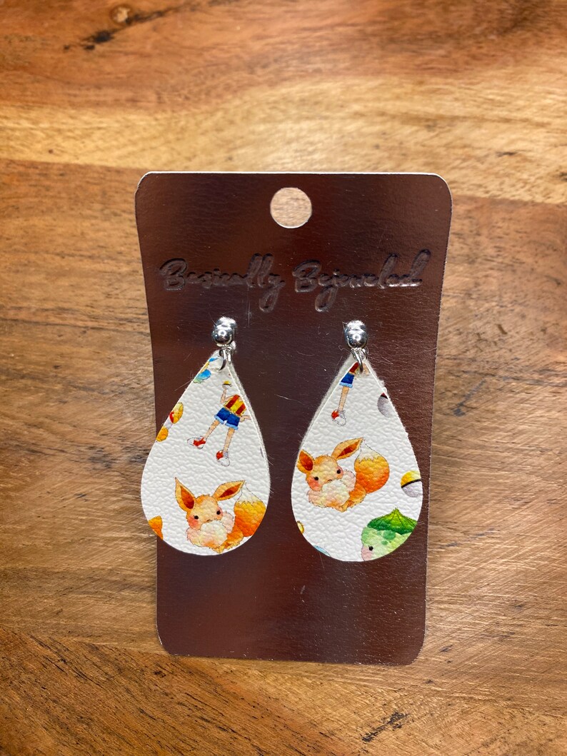 Puede incluir: Un par de pendientes blancos en forma de l&aacute;grima con un estampado de personaje de Pokemon. Los pendientes presentan un peque&ntilde;o Pokemon naranja parecido a un zorro sobre un fondo blanco. Los pendientes est&aacute;n en una tarjeta plateada con el texto "Basically Bejeweled".