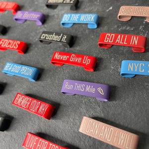 May include: A collection of colourful, motivational keychains with various phrases, including "Be Better", "Do The Work", "Go All In", "Never Give Up", "Run This Mile", and "Remember Your Why".