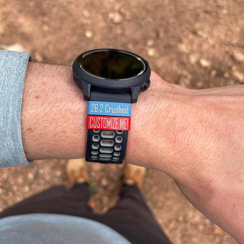 May include: A black smartwatch with a black and white patterned strap. The strap has two colourful bands on top of it, one blue with the text "26.2 Crushed" and one red with the text "CUSTOMIZE ME!"