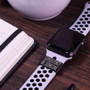 May include: A black and white Apple Watch with a black band that says "Consistency Be Someone".