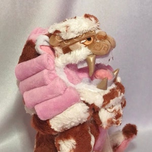 Puede incluir: Un juguete de peluche de tigre con pelaje marrón y blanco, detalles rosas y detalles dorados. El tigre está sentado con la boca abierta, mostrando sus dientes.