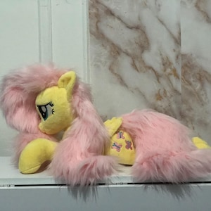 May include: A plush toy of a yellow pony with long, shaggy pink hair and a pink tail. The pony has large blue eyes with long eyelashes and a cutie mark on its flank featuring three small pink butterflies. The toy is in a lying position.