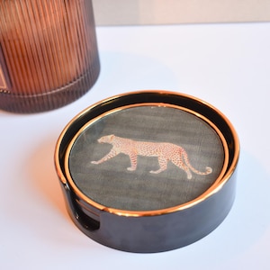 May include: A set of black ceramic coasters with gold rims, featuring a leopard print design. The coasters have a dark grey background with an orange and brown leopard. The set is a stylish addition to any home.