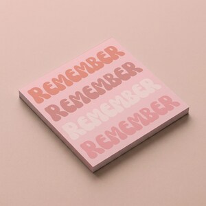 May include: A square pink notepad with the word "REMEMBER" repeated in a retro font. The word is in shades of peach, pink, and white. The notepad is on a matching pink surface.