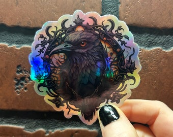 May include: A holographic sticker featuring a detailed illustration of a raven's head with a crown, surrounded by intricate vines and flowers.