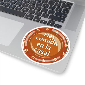 May include: A round sticker with the text "Hay comida en la casa!" in white on a brown background. The sticker has a decorative border with floral and leaf patterns. The sticker is placed on a laptop.