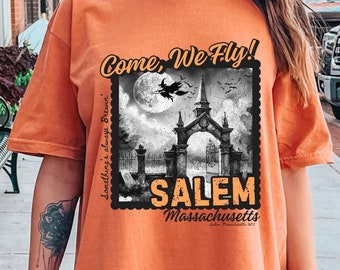 May include: Orange t-shirt with a graphic print of a spooky silhouette of a church with a witch flying over it. The text "Come, We Fly!" is above the image and "Salem, Massachusetts" is below the image.