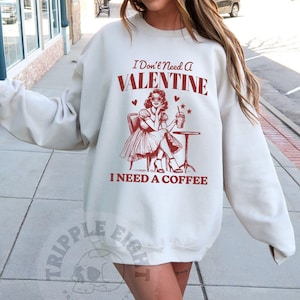 May include: A white sweatshirt with a red graphic print of a woman sitting at a table with a coffee cup. The text on the sweatshirt reads "I Don't Need A Valentine I Need A Coffee".