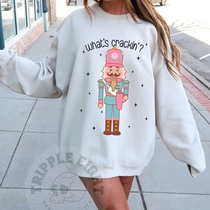 Może przedstawiać: Piaskowa bluza z nadrukiem żołnierza-dziadka do orzechów. Dziadek do orzechów jest w odcieniach różu, niebieskiego i brązu, z napisem "what's crackin'?" powyżej. Bluza ma luźny krój.