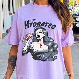 May include: A lavender t-shirt with a graphic of a vampire woman drinking from a glass of red liquid. The text "STAY HYDRATED" is above the graphic.