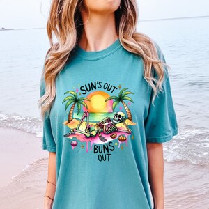 Op de afbeelding: Een turquoise t-shirt met een afbeelding van een skelet dat op een strand ligt met palmbomen en de tekst "SUN'S OUT BUNS OUT".
