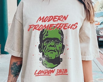 Puede incluir: Camiseta color marfil con un gráfico verde y negro del monstruo de Frankenstein y el texto rojo "MODERN PROMETHEUS LONDON 1818". La camiseta tiene mangas cortas y un corte holgado.