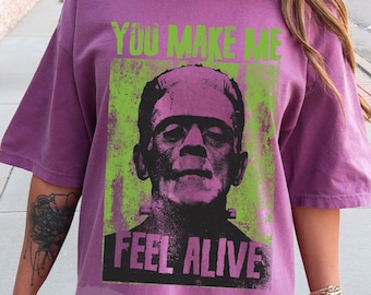Puede incluir: Camiseta morada con un gráfico verde de Frankenstein y el texto "You Make Me Feel Alive".
