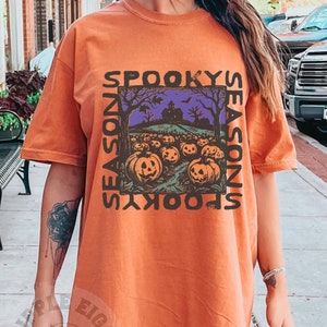 May include: Orange t-shirt with a graphic print of a spooky season scene with pumpkins and a house. The text "Spooky Season" is written vertically on either side of the graphic.