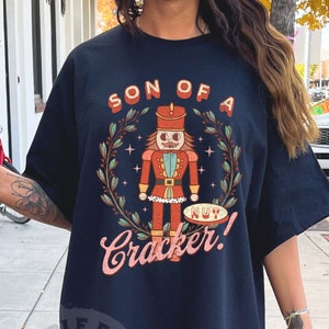 May include: Black t-shirt with a graphic of a nutcracker holding a sign that says "Nut Cracker!" and the text "Son of a" above the nutcracker.