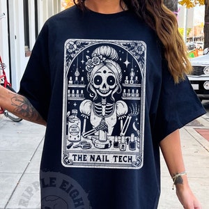 May include: Black t-shirt with a white graphic of a skeleton woman with a skull face, holding a bottle of nail polish and a pair of nail clippers. The graphic is in a tarot card style frame with the text "The Nail Tech".
