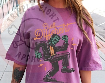 Puede incluir: Camiseta color bayas con un gráfico de Frankenstein bailando. El texto "Built Different" está encima del gráfico en una fuente amarilla. La camiseta tiene mangas cortas y un corte holgado.