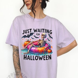 May include: A lavender t-shirt with the text "Just Waiting for Halloween" and a skeleton wearing a colorful inflatable ring. The skeleton is surrounded by bats, pumpkins, and candy.