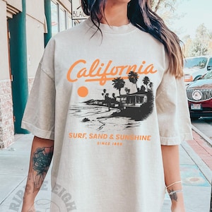 May include: A light beige oversized t-shirt with a graphic print of a beach scene and the text "California Surf, Sand & Sunshine Since 1959".