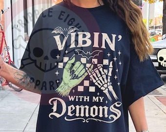 Puede incluir: Camiseta negra con el texto "VIBIN' WITH MY DEMONS" y un gráfico de una mano verde y una mano de esqueleto. El diseño incluye murciélagos y un patrón de tablero de ajedrez. La camiseta es de corte holgado.