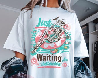 May include: White t-shirt with a Halloween graphic. The design shows a skeleton on a pool float with the text "Just Waiting for Halloween". The graphic includes a bat, sweets, and pumpkins.