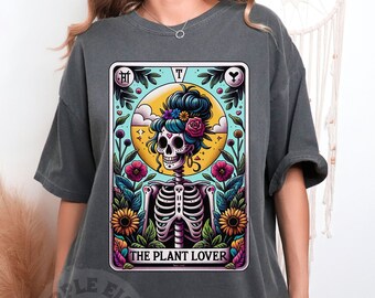 Puede incluir: Camiseta gris oscuro con un diseño de carta del tarot que presenta un esqueleto adornado con flores y el texto "THE PLANT LOVER". El diseño incluye un fondo floral y celestial colorido.