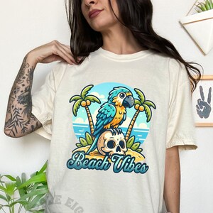 May include: A white t-shirt with a blue and yellow parrot perched on a skull with palm trees and ocean waves in the background. The text "Beach Vibes" is printed below the parrot.