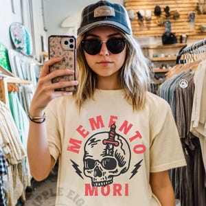 Puede incluir: Una mujer lleva una camiseta beige claro con un gr&aacute;fico de calavera y el texto "Memento Mori" en rojo. La calavera tiene una daga que la atraviesa. La camiseta es de estilo unisex.
