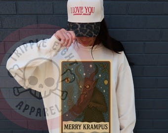 May include: Cream-colored sweatshirt with a Krampus graphic and the text "MERRY KRAMPUS". The image also shows a white baseball cap with the text "I LOVE YOU" in red.