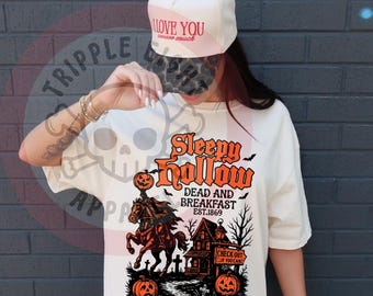 May include: Ivory-colored t-shirt with a Halloween-themed graphic. The design features the text "Sleepy Hollow Dead and Breakfast EST.1869" with a headless horseman, pumpkins, and a haunted house. The shirt also has the text "The place to rest a weary head."