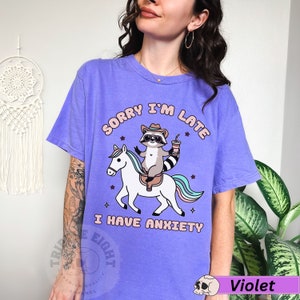 May include: Purple t-shirt with a graphic of a raccoon riding a unicorn with the text "Sorry I'm Late I Have Anxiety".