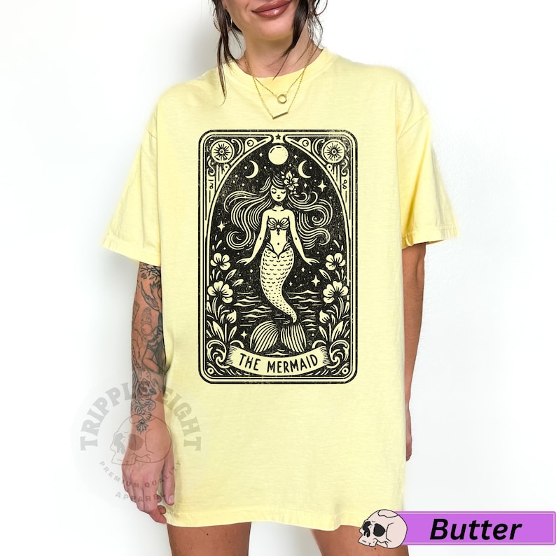 May include: A yellow t-shirt featuring a black and white illustration of a mermaid within a tarot card style frame. The text "The Mermaid" is printed beneath the image.