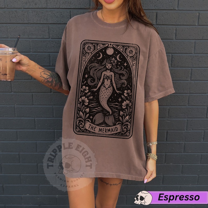 May include: A brown t-shirt featuring a black graphic of a mermaid in a tarot card style design. The text "The Mermaid" is printed below the image. The shirt has a relaxed fit and is made of a soft, comfortable fabric.