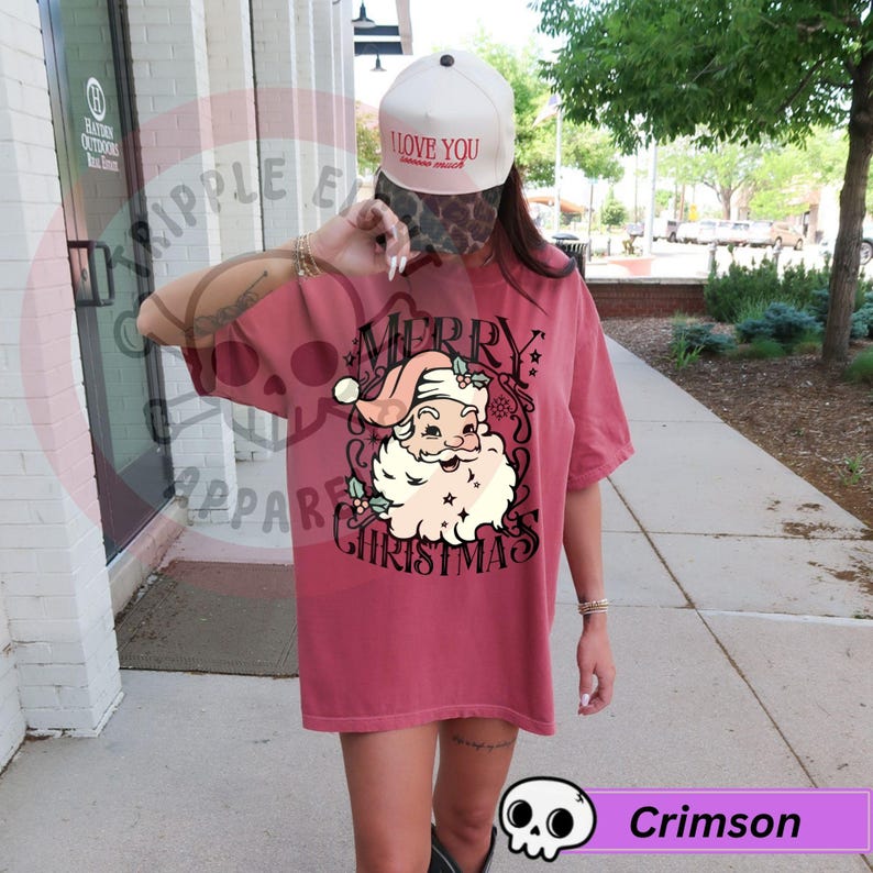 May include: Crimson-colored t-shirt featuring a Santa Claus graphic with the words "Merry Christmas". The Santa illustration has a white beard and a pink hat. The shirt is a short-sleeved, oversized style.