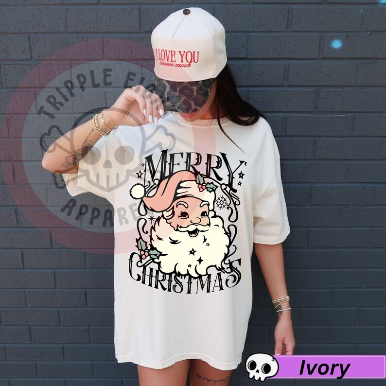 May include: Ivory-colored t-shirt with a vintage Santa Claus graphic. The design features Santa in a pink hat and beard, with the words "MERRY CHRISTMAS" in a decorative font. A white baseball cap with the text "I LOVE YOU" is also visible.