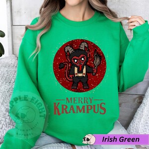 May include: Green Christmas sweatshirt with a red circle graphic of a cartoon Krampus holding a stick. The text "Merry Krampus" is printed below the graphic.