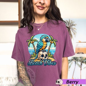 May include: A purple t-shirt with a cartoon illustration of a blue and yellow parrot perched on a skull. The parrot is surrounded by palm trees and the text "Beach Vibes" is printed below the image.